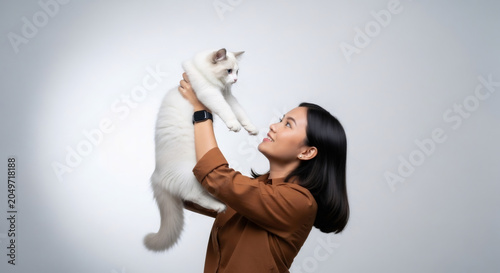A happy young Asian woman in a brown shirt affectionately holds up her white cat in a bright studio setting.