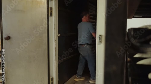 Male baker moving a large baking rack loaded with trays of raw bread dough out of a leavening chamber in a professional bakery kitchen