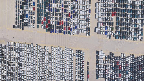 Aerial drone view moving over thousands of new manufactured cars neatly parked and stored in a large industrial parking lot, waiting for distribution, export, and sale in the automotive market
