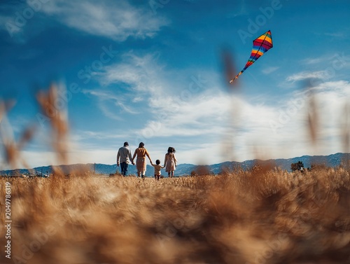 Family walks through dry grass field, flying colorful kite under blue sky