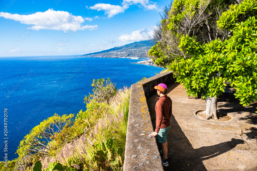 Obraz premium Female tourist stands at a walled viewpoint looking at the blue coast, La Palma, Canary Islands, Spain