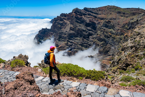 Fotografie Hiker in a bright orange jacket and pink cap looks out over a massive, rugged ca