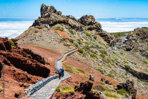 Two hikers walk down a paved mountain path leading toward a jagged volcanic peak above a sea of clouds near Los Andenes, La Palma, Canary Islands, Spain