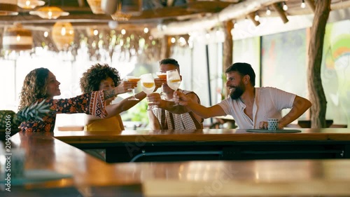 Group of four happy, diverse friends sitting at a wooden bar counter in a tropical themed bar, toasting and celebrating together