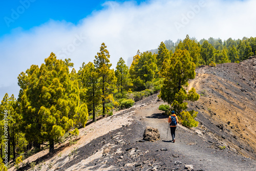 Hiker with a blue backpack walks along a high volcanic ridge path lined with vibrant green pine trees on Ruta de los Volcanes, La Palma, Canary Islands, Spain