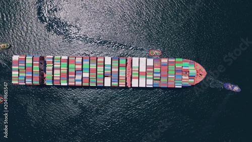 Aerial top-down view of a massive container ship loaded with colorful cargo containers being maneuvered by several powerful tugboats on the open ocean for international trade and shipping