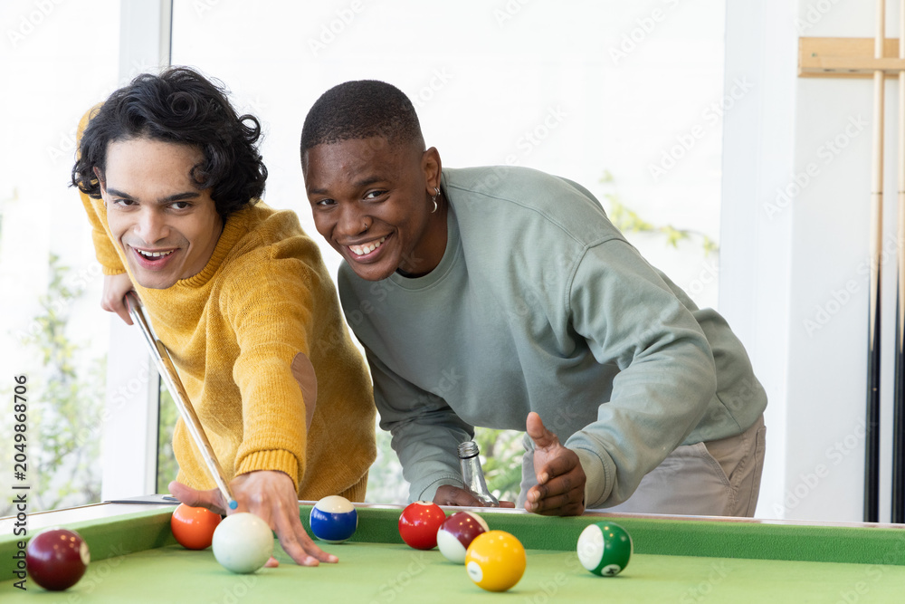 Obraz premium Diverse male friends in mustard sweater, light sweatshirt playing on green-felt table in game room