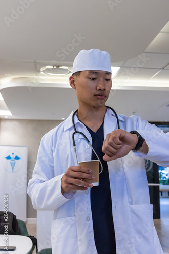 Asian male doctor in lab coat with stethoscope checking watch while holding coffee cup at hospital