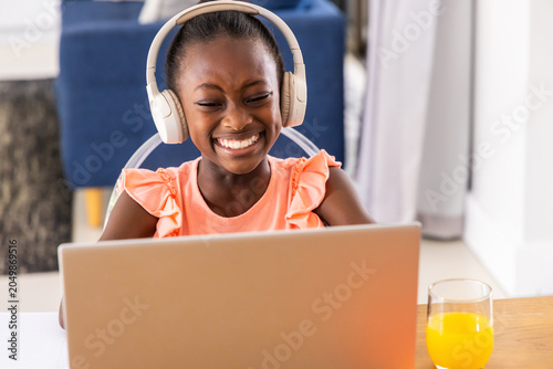 Female child smiling, wearing white headphones, using laptop at home on table with orange juice