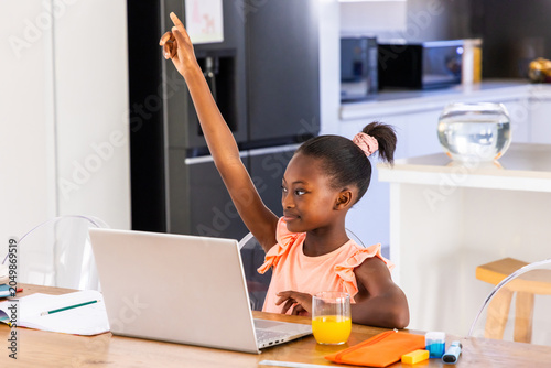 African American youth female raising arm studying on laptop in home kitchen with orange juice