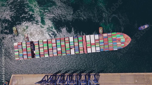 Impressive top-down aerial footage capturing a massive cargo vessel loaded with colorful containers being expertly maneuvered into a commercial harbor by a fleet of assisting tugboats