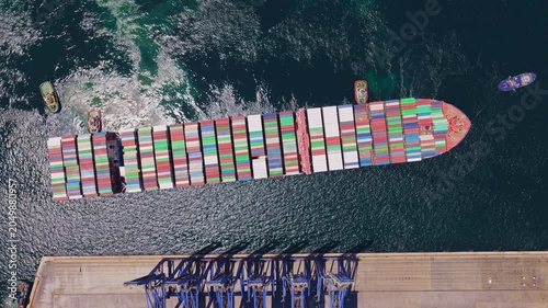 Huge container ship filled with colorful cargo boxes departing from a commercial port terminal, assisted by several small tugboats maneuvering it into the open sea for international export