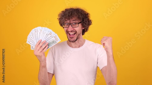 Excited young man celebrating financial success holding cash fan, raising clenched fist, bright yellow backdrop boosting joyful emotion, strong commercial appeal for money, achievement and lifestyle