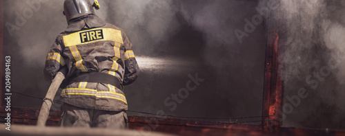 Firefighter spraying water on burning building, heavy smoke and flames, emergency firefighting action, back view of fireman in protective gear during dangerous fire rescue operation