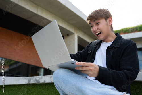 Young Asian graduate celebrating success with laptop outdoors in front of modern building, symbolizing achievement, startup growth, and positive career lifestyle with joyful expression, technology