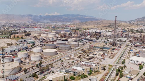 Expansive aerial perspective of an oil refinery and storage facility, showcasing the intricate network of pipelines, smokestacks, and large white tanks under a partly cloudy sky