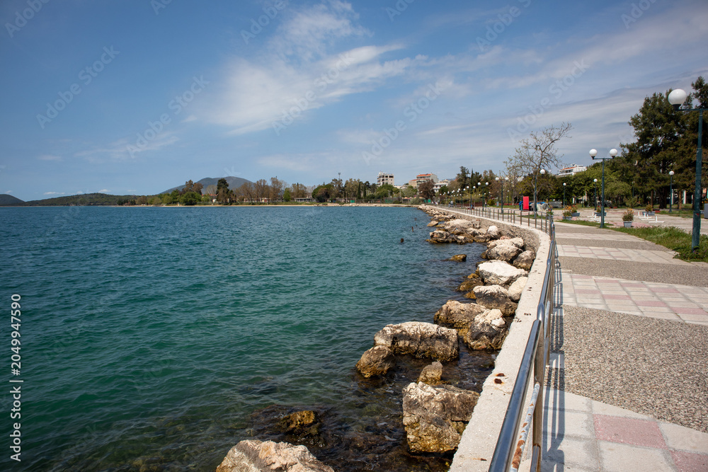 Fototapeta premium Seaside promenade with rocky shoreline and calm sea water under blue sky in Igoumenitsa, Greece.