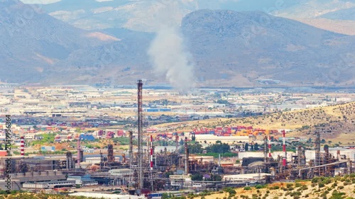 Large smoke plume rising from a fire that broke out directly behind an operational oil refinery. Distillation towers in the foreground and arid mountains in the background.