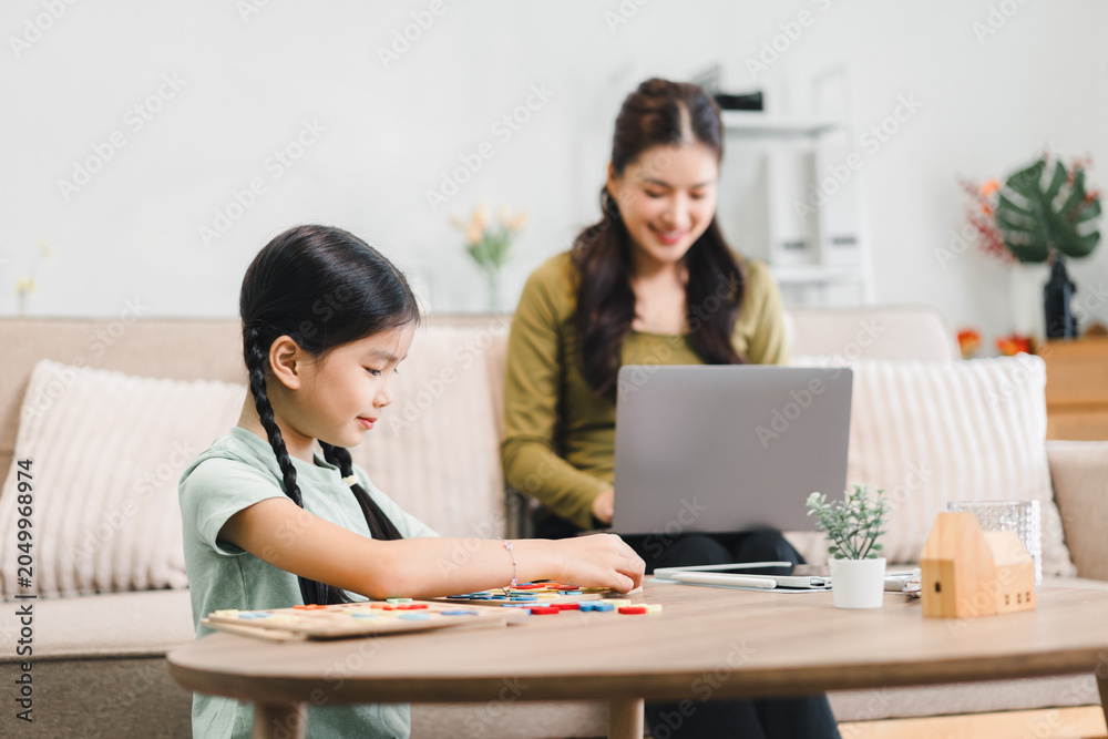 Obraz premium Asian mother working on a laptop while her daughter plays with educational toys at home