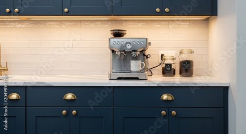 A modern home coffee station featuring a silver espresso machine with a white cup on a white marble countertop against dark blue kitchen cabinets and warm under-cabinet lighting.