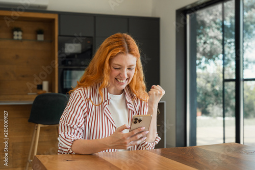 Canvas-taulu Happy redhead woman at home celebrating success and good news on her smartphone,