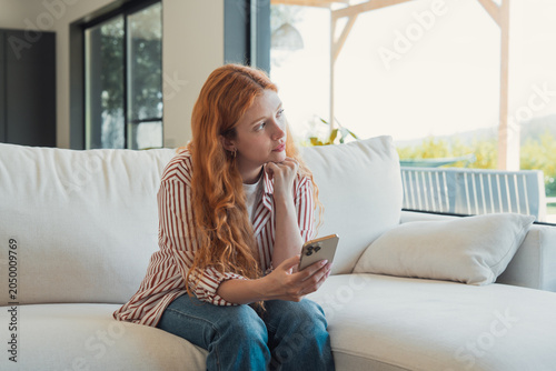 Konstfotografi Young woman with red hair sitting on a white sofa in a modern living room, holdi