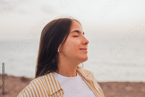 Young woman with eyes closed meditating on a tranquil beach at sunset, breathing deeply and feeling calm, serene and mindful by the ocean shoreline