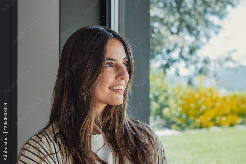 Obraz premium Woman with long brunette hair smiling while looking away through a window, experiencing emotions of hope, peace, serenity, and a sense of well-being indoors