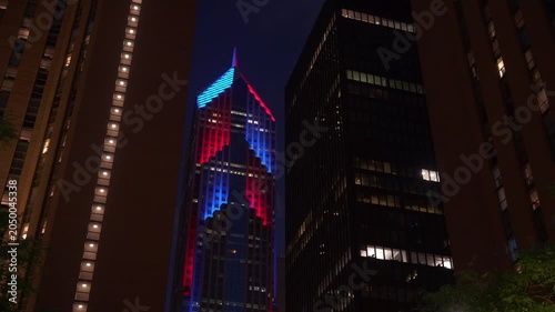 Tall Chicago skyscraper glowing with red and blue lights between dark high-rise buildings at night. Dramatic urban cityscape with illuminated windows and modern architecture.