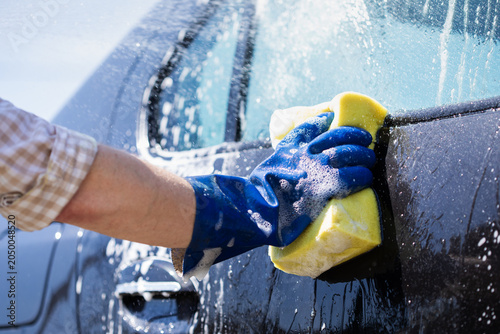 Car wash with worker hand holding sponge, washing off auto shampoo soap foam and cleaning transport vehicle on carwash sunny outdoors with water rinse.