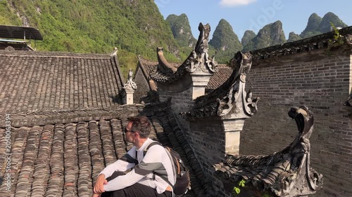 Slow motion shot of a male traveler with a backpack sitting on a traditional undulating tiled roof of a Ming‑and‑Qing‑era house, overlooking a scenic karst mountain landscape in Xingping Fishing Villa
