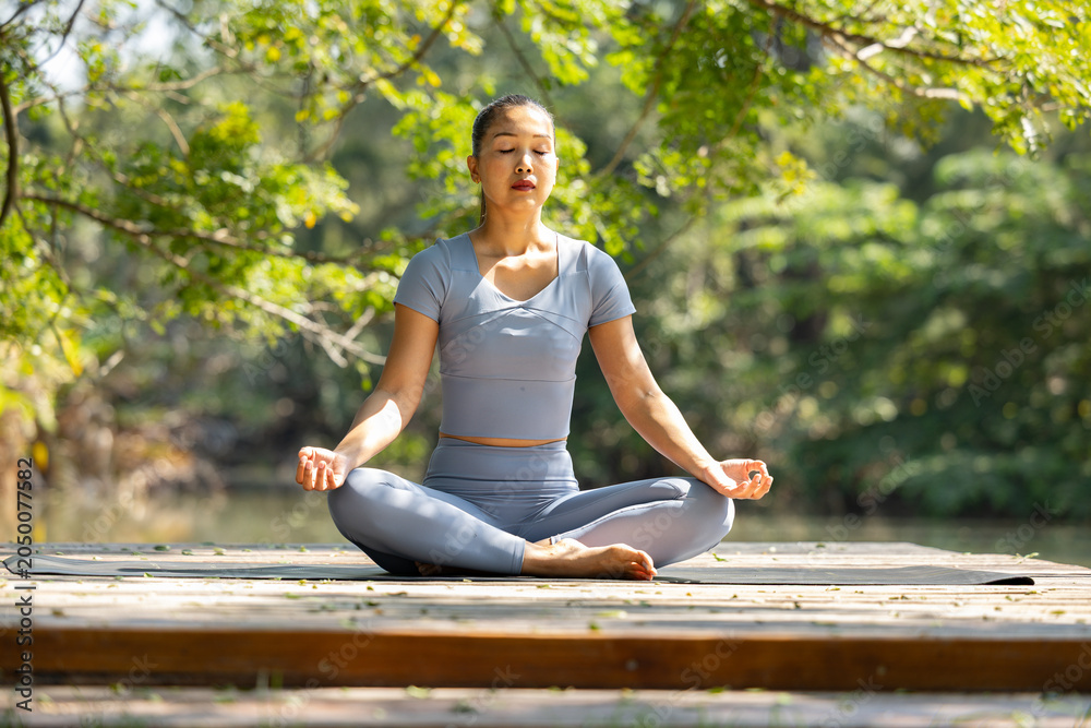 Fototapeta premium Yoga woman meditation outdoor nature sitting calm exercise woman practicing yoga meditation sitting outdoor on wooden platform surrounded by green forest sunlight