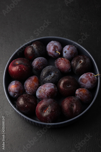 Juicy ripe fresh plum with water drops in gray plate on black background. Seasonal fruits. Selective focus, close-up.
