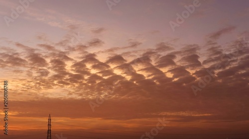 Atmospheric Sunset Sky Background with Dramatic Rippled Cloud Formations and Golden Hour Hues Featuring a Silhouetted Structure at Horizon