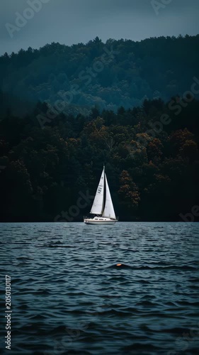 sailboat with white sails crossing calm dark waters before forested hills under overcast sky