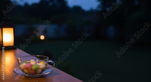 A cup of kopi tubruk sits on a wooden railing at dusk with colorful sweets inside and a lantern nearby in a serene outdoor setting.
