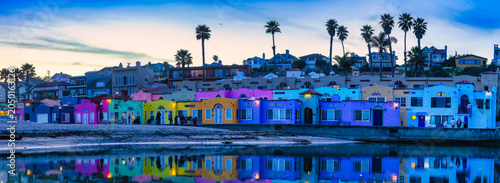 Wide panoramic view of colorful beachfront houses at Capitola Beach with reflections in calm water at sunset, palm trees and coastal neighborhood along California Pacific coast