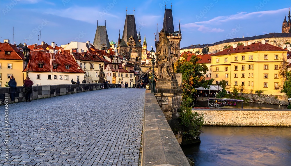 Fototapeta premium A cobblestone bridge lined with statues leads to a historic cityscape under a blue sky with scattered clouds