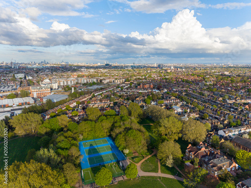 LONDON, UK - JUNE 9, 2026: Drone high-angle panoramic view of residential areas, a suburban park with blue tennis courts, and the city skyline under a cloudy summer sky