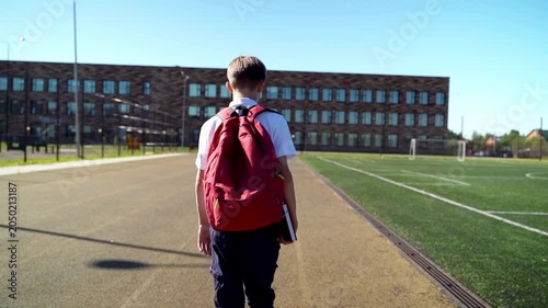 Boy walks toward school building. Red backpack sits on his shoulders. He carries books, moving slowly. Clear sky, bright sun, empty field behind him. Ideal for school, education