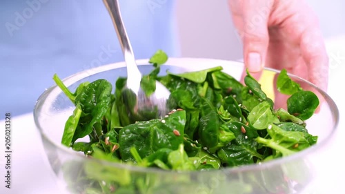 Hand mixes fresh greens in a clear bowl. Bright lighting highlights crisp, leafy vegetables. No visible emotion from the hand. Ideal for healthy eating or kitchen prep scenes