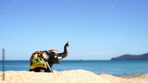 Small elephant statue on sandy beach. Blue sky and ocean behind it. Coastal landscape with distant hills. Still scene evokes calm and vacation. Great for travel, holiday, or tropical themes