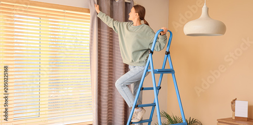 Cuadro en lienzo Young woman on stepladder hanging curtain at home