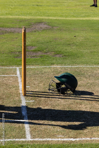 Cricket helmet is lying on worn brown pitch near stump with white creases and shadows