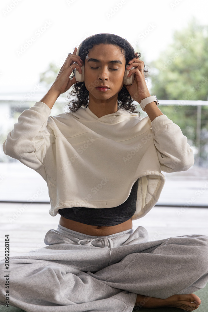 Obraz premium Female meditating on yoga mat by glass door holding headphones, wearing hoodie and smartwatch
