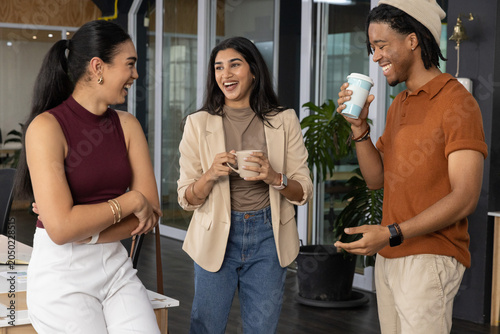 Three coworkers chatting in office wearing blazer, holding mug and travel cup near glass doors