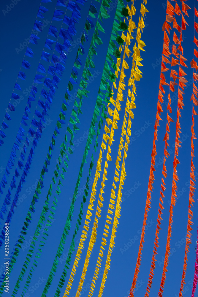 Fototapeta premium Lindo contraste de varais repletos de bandeirinhas juninas coloridas sob céu azul limpo. Imagem vertical.