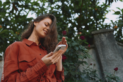 Tableau sur toile A smiling woman in a rust shirt checks her phone outdoors, with lush greenery in