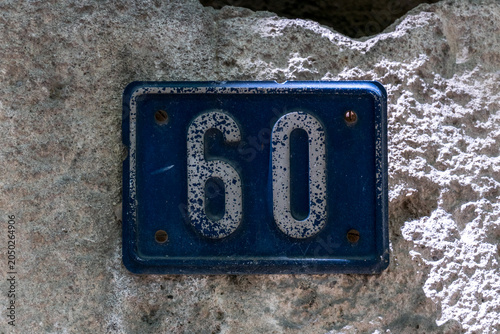 A ceramic house number 60 plaque with blue numerals on a cream tile, mounted on a weathered stone wall in a French village