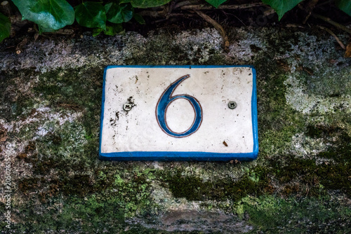 A ceramic house number 6 plaque with blue numerals on a cream tile, mounted on a weathered stone wall in a French village
